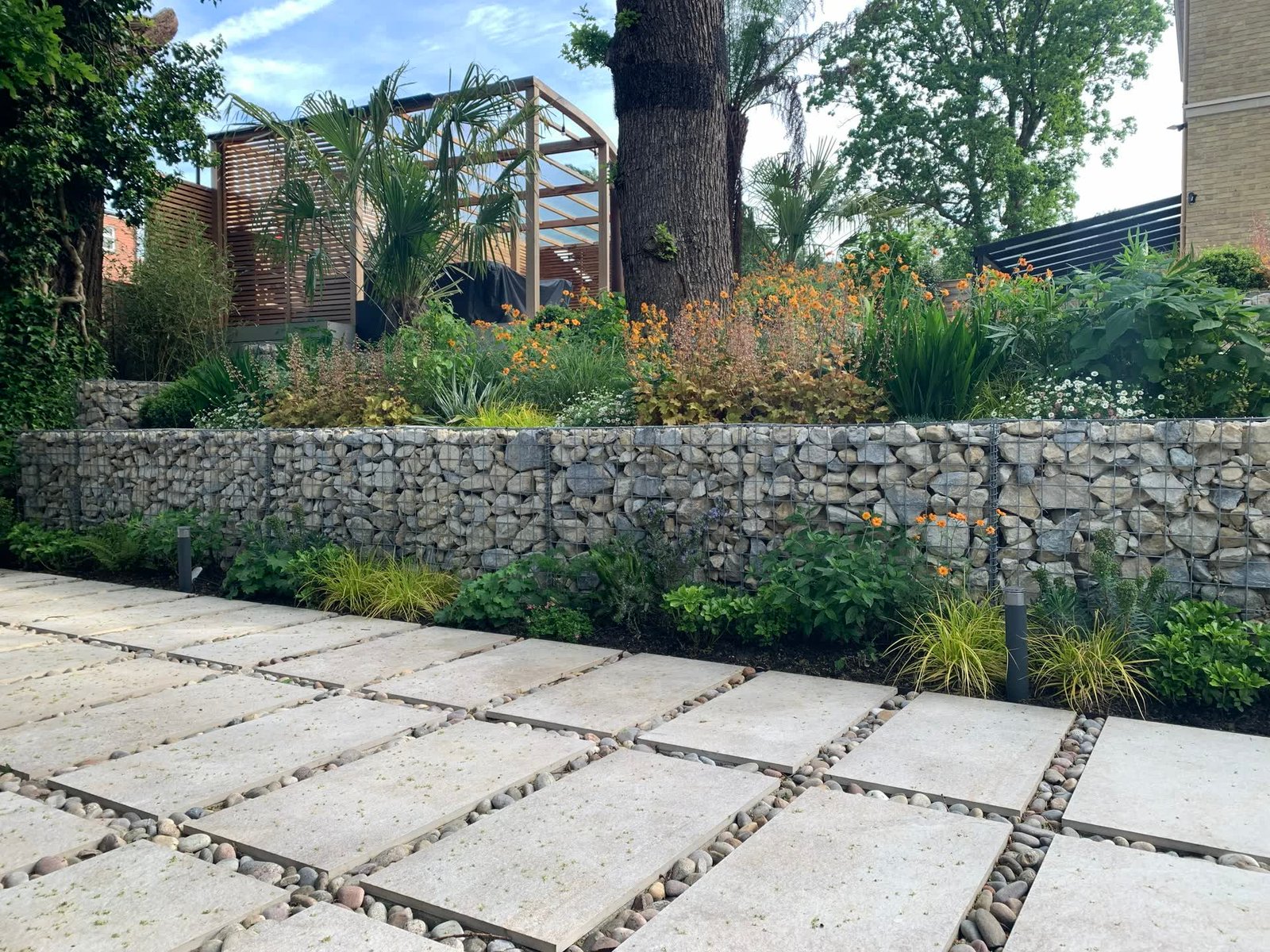 Gabion retaining wall holding a raised bed of grasses and late-summer perennials, above large-format textured paving with pebble-filled joints