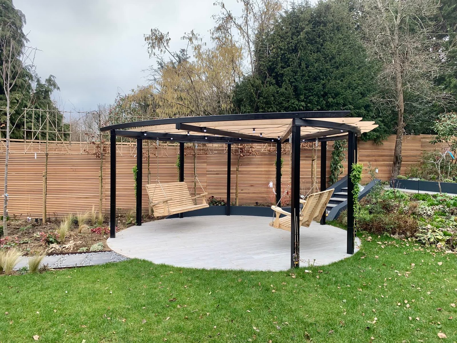 Curved black steel and timber pergola with two hanging swing seats, over a circular pale porcelain terrace set into a large lawn