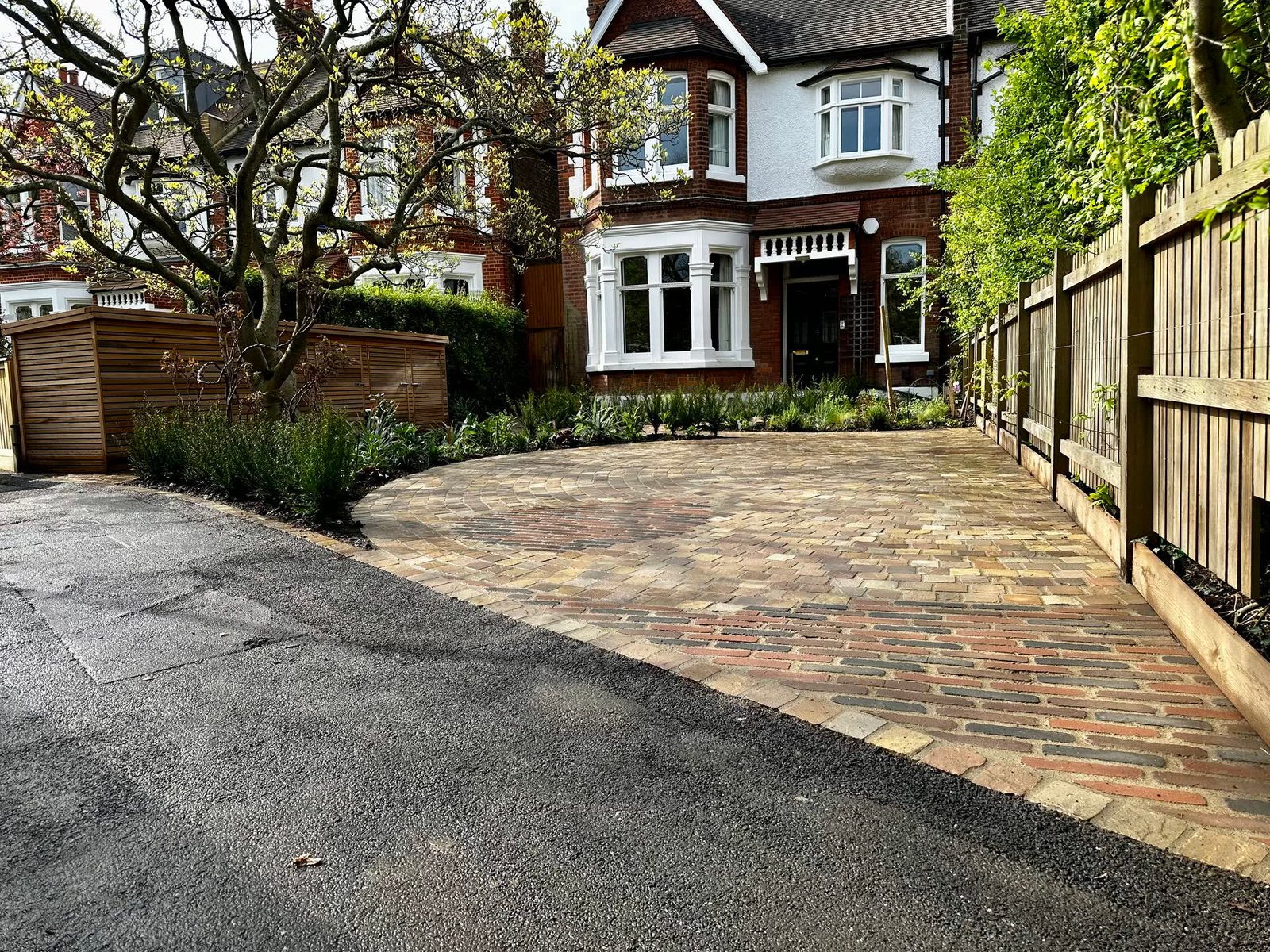 Reclaimed stone sett driveway laid in a fan pattern, edged with a planted border and a slatted timber bin store, in front of a bay-fronted Edwardian semi